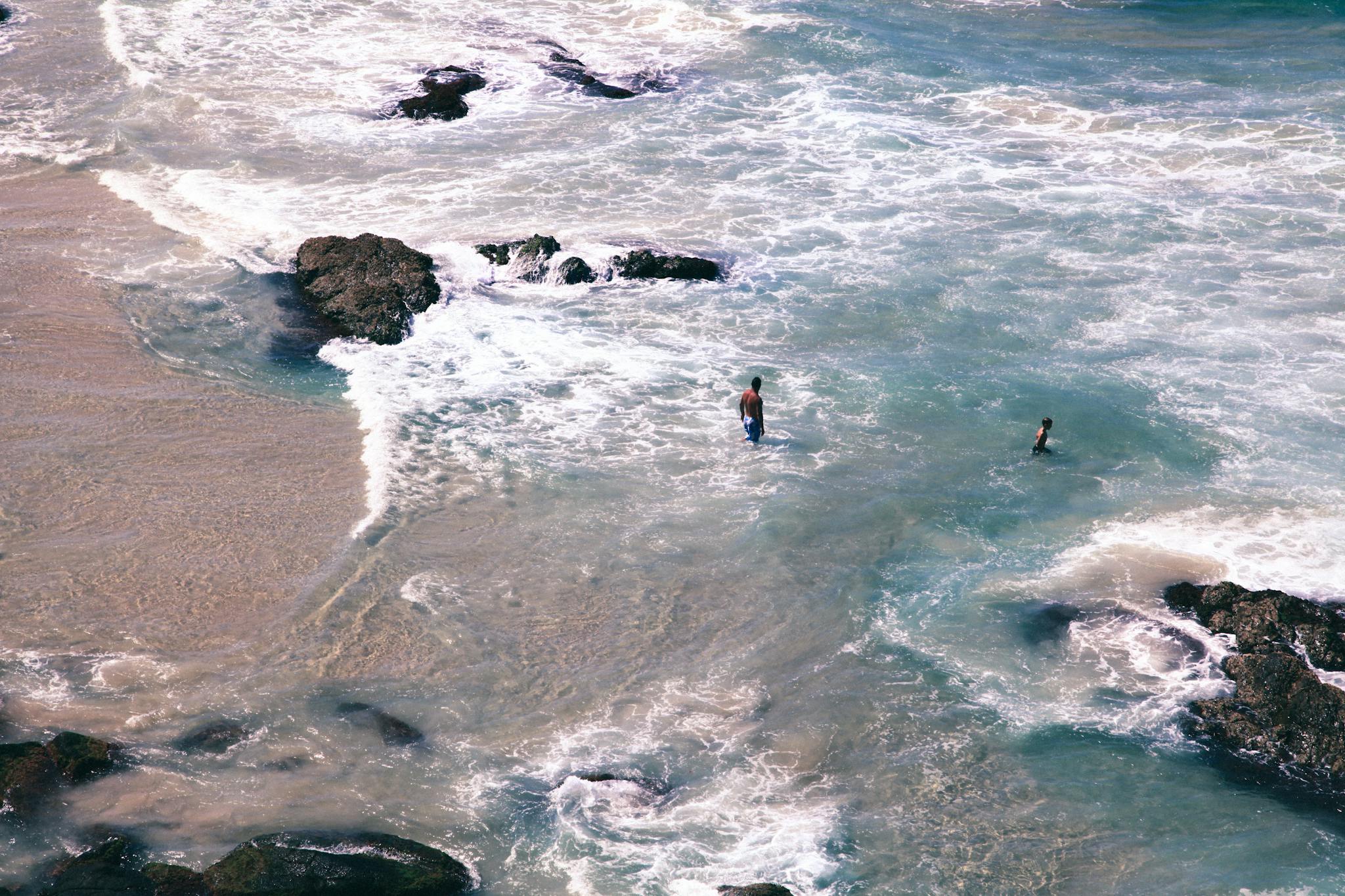 Aerial shot capturing people swimming and walking on a rocky beach with waves crashing.